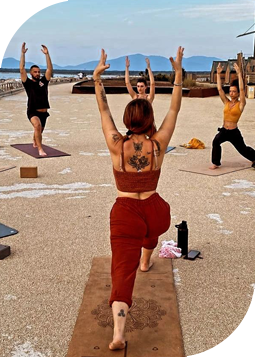 corso-anukalana-yoga-pisa Gruppo di persone che pratica Anukalana Yoga all'aperto su una terrazza con vista mare, eseguendo movimenti fluidi in una posa di affondo con le braccia sollevate